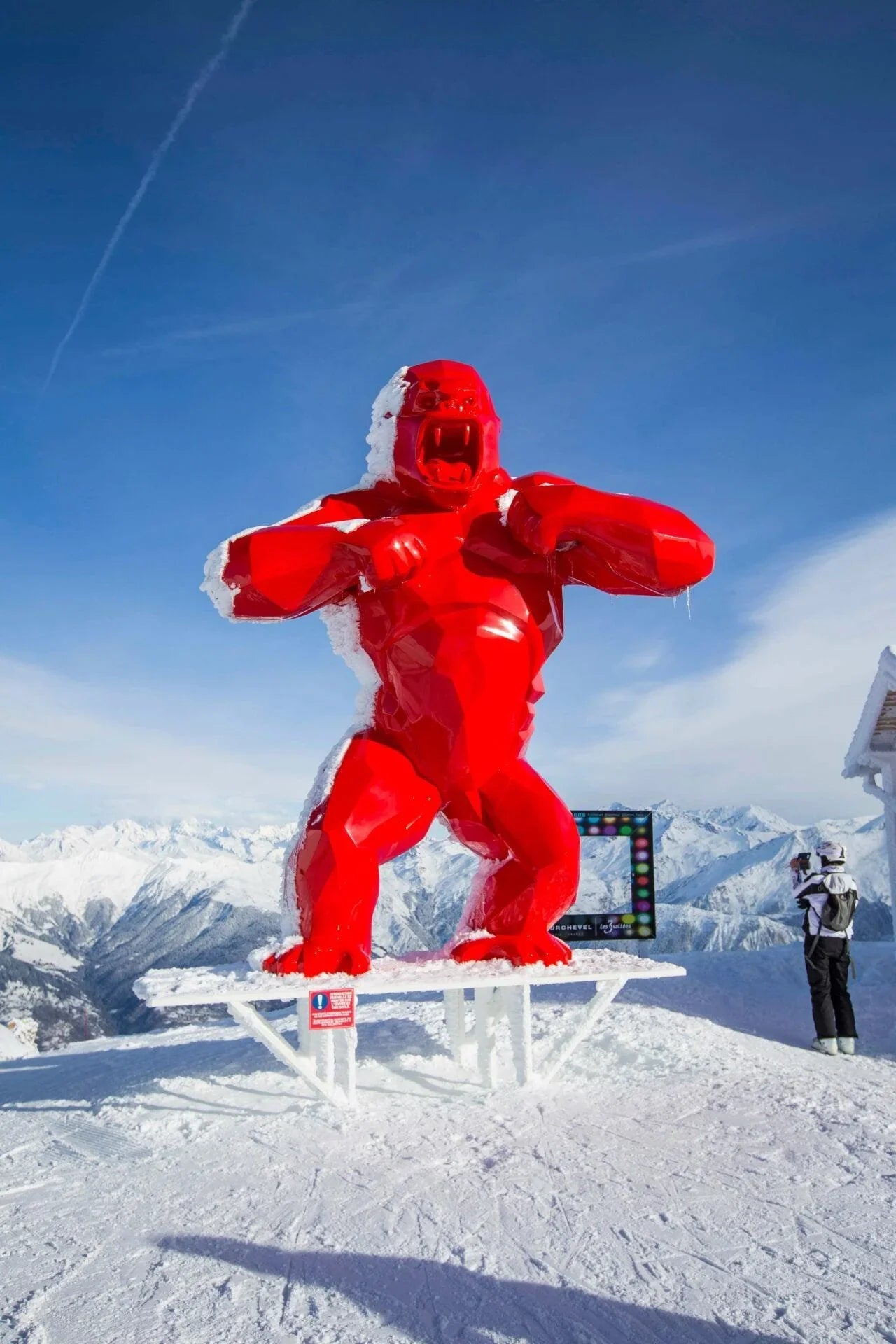 Bright red angular gorilla sculpture covered with snow on a snowy mountain peak under a clear blue sky